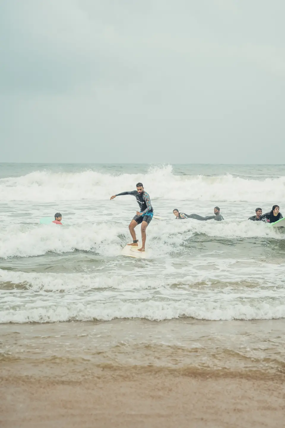 A man in a dark rash guard rides whitewater on a cream board while others lie on their boards behind him