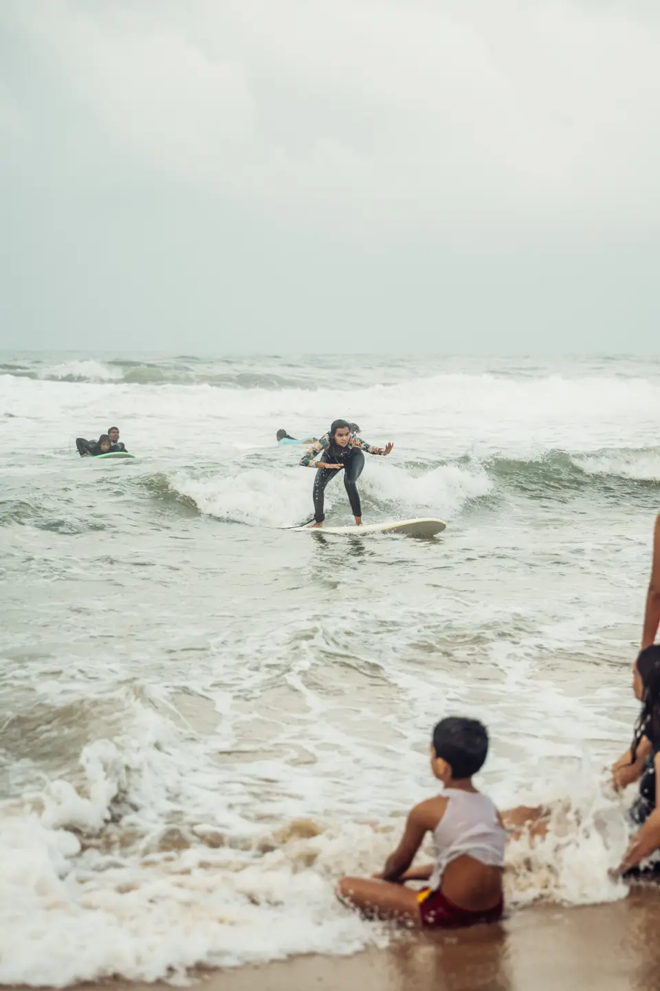 A learner standing up on a board in the whitewater while a small child sits in the foam at the water's edge