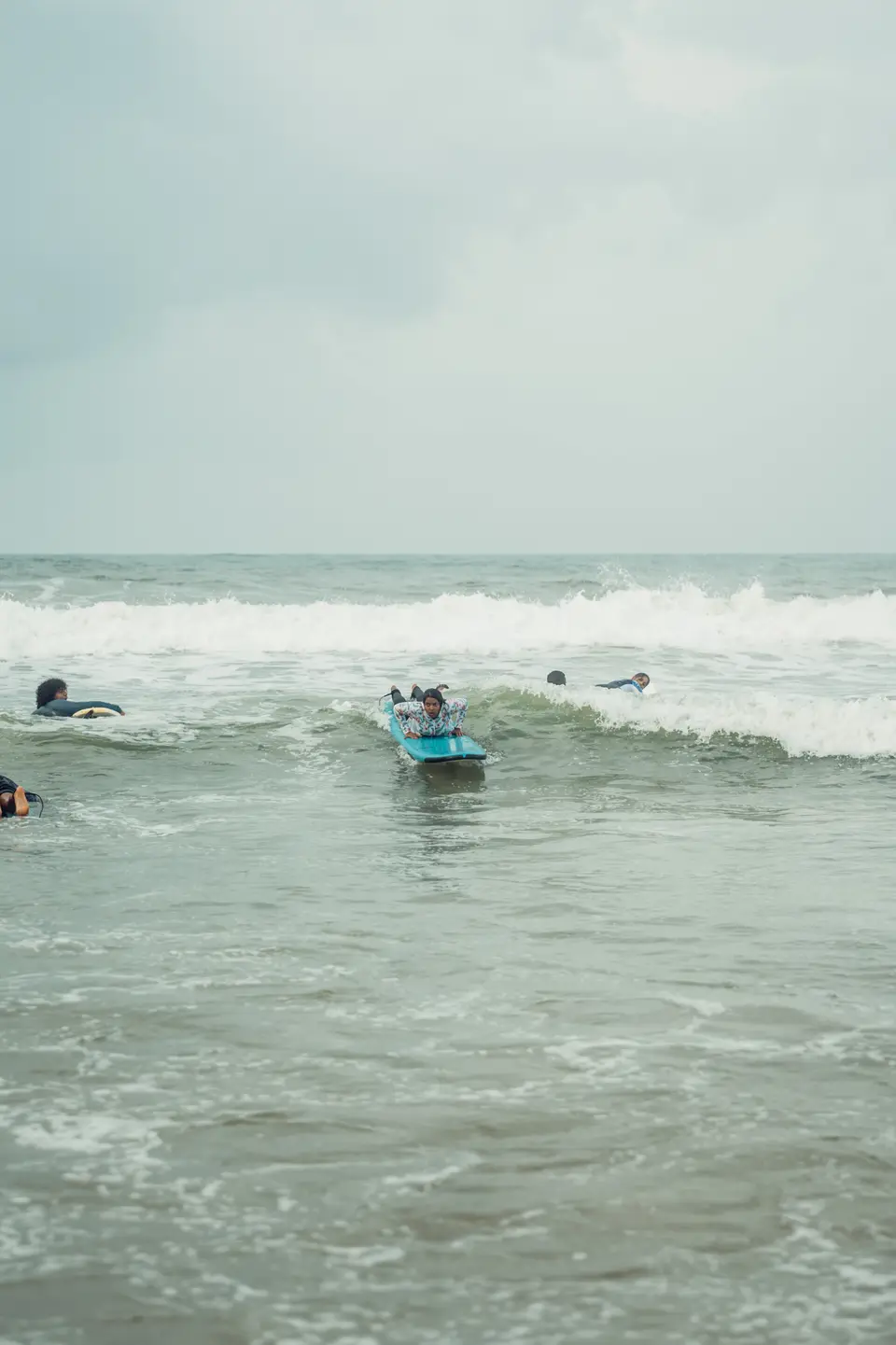 A woman in a floral top lies flat on a blue soft-top as foam carries her in, others on boards nearby