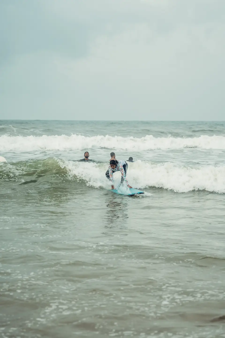 A learner pushing up to stand on a blue board as a small wave breaks, three people in the water behind