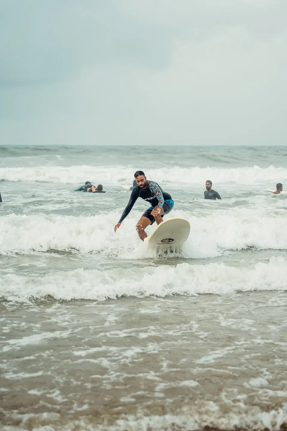 A man in a black rash guard with patterned sleeves crouches on a cream soft-top as its nose lifts out of the foam
