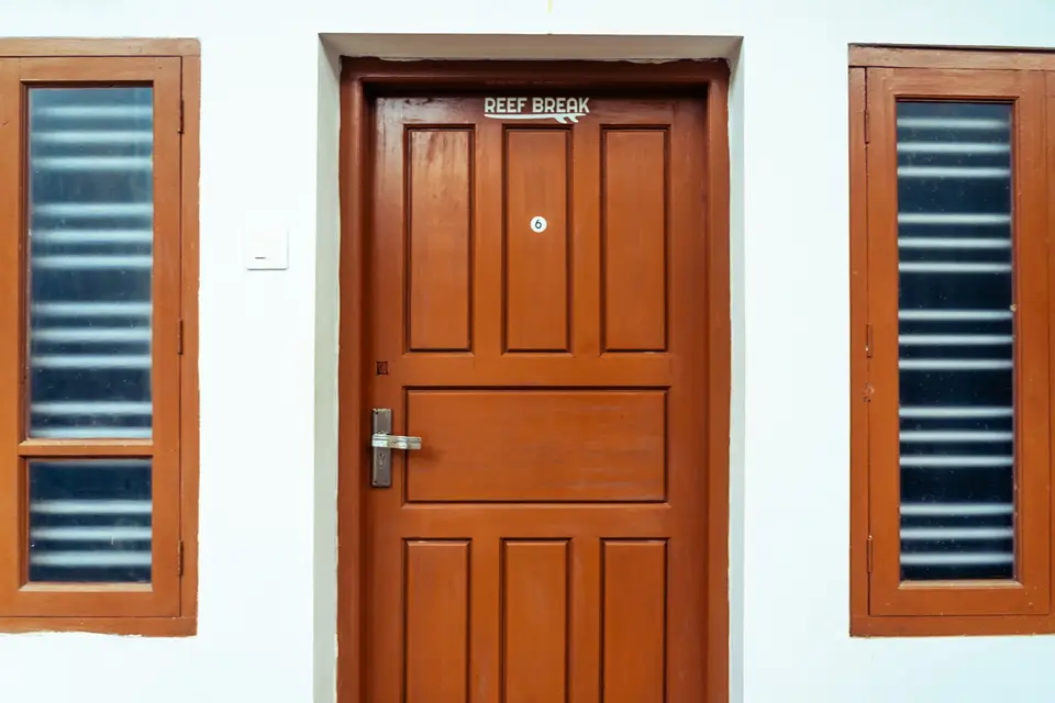 A wooden door signed Reef Break in a white wall, between two windows with frosted glass slats