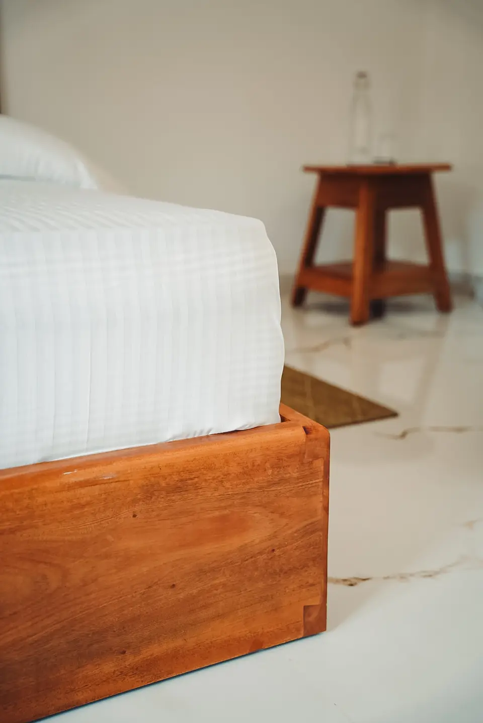 Corner of a wooden bed base with an interlocking finger joint, white bedding above and marble floor below