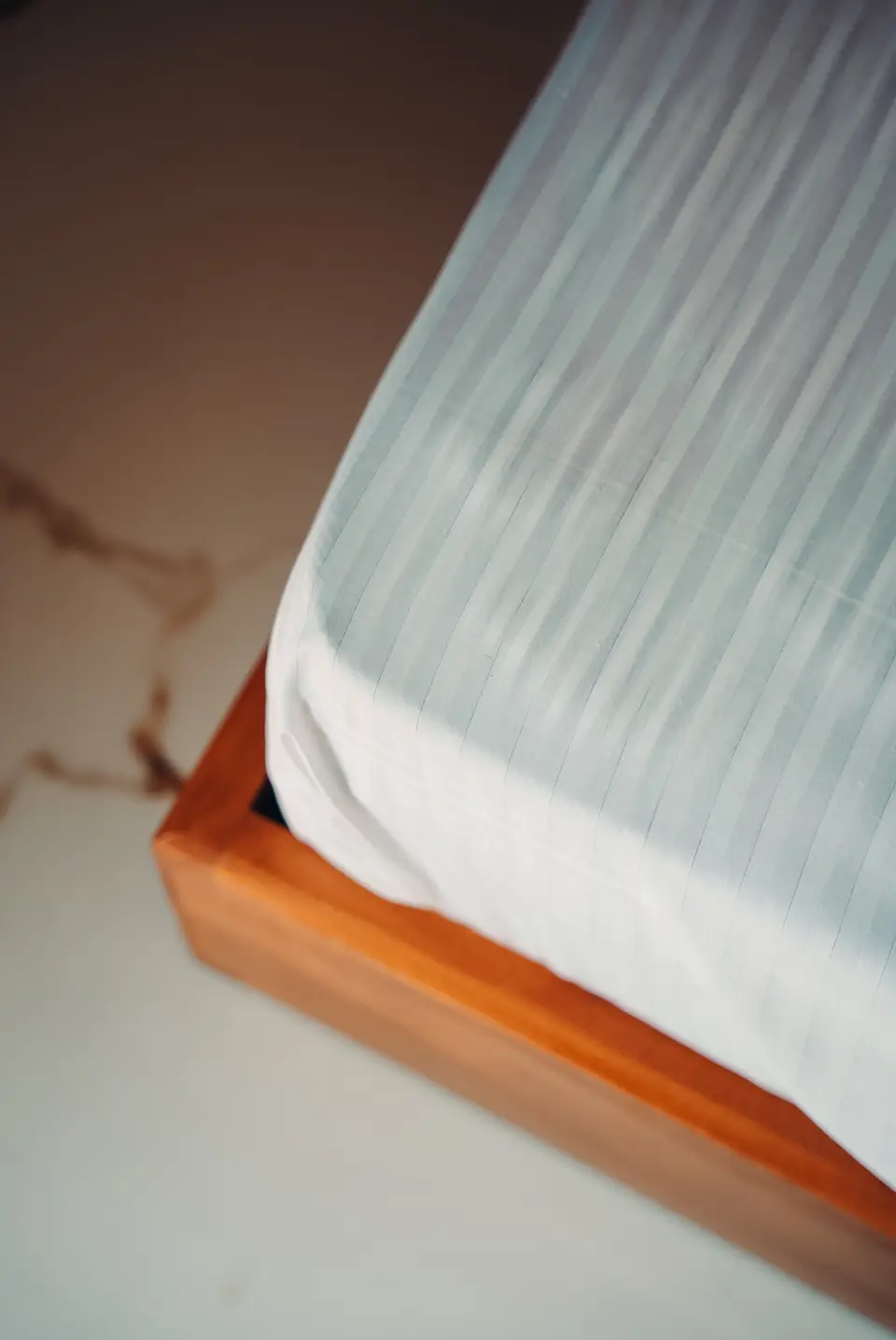 Corner of a mattress with a striped white sheet, sitting inside a wooden bed frame on a marble floor