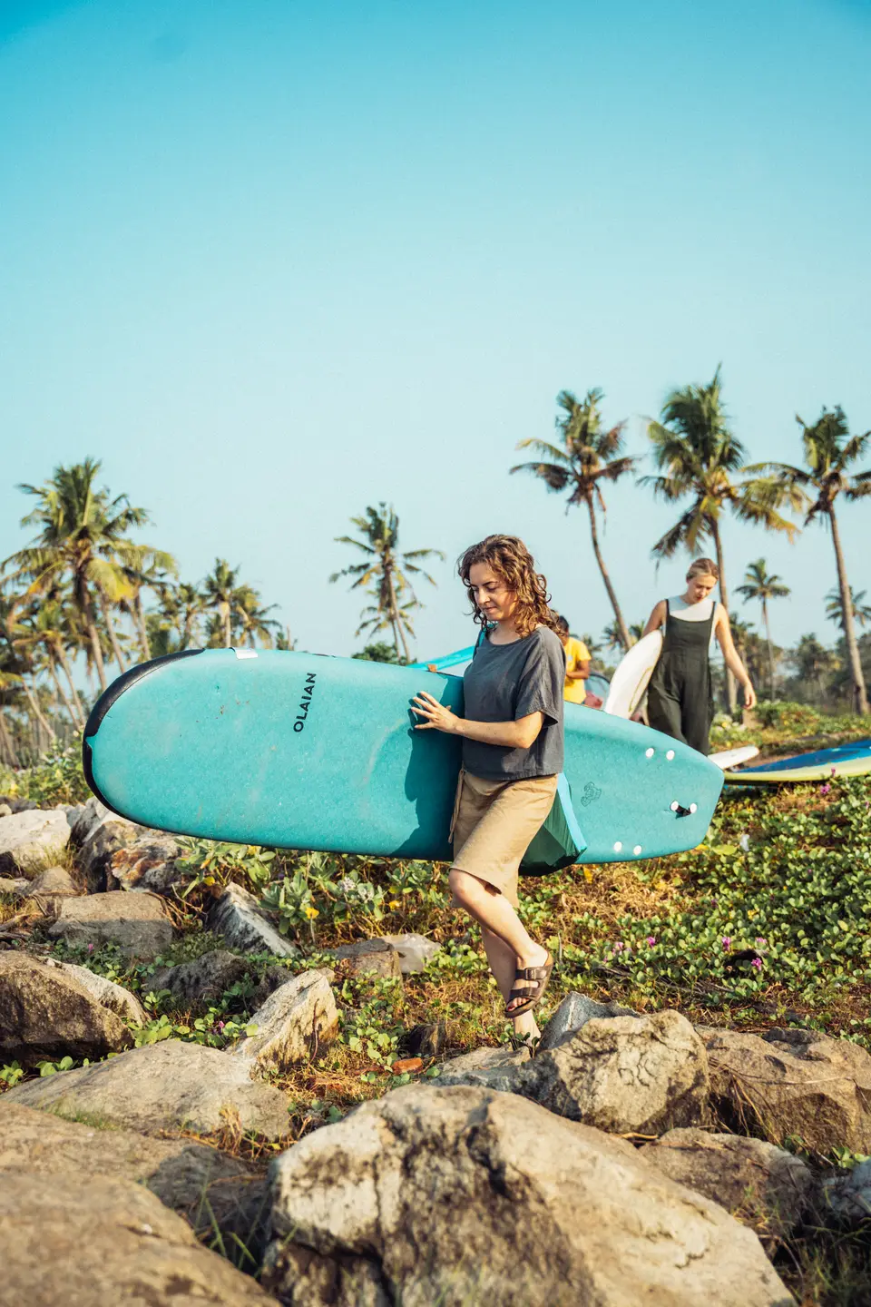 A woman in sandals carries a teal soft-top over coastal rocks and green creepers, another woman with a white board behind