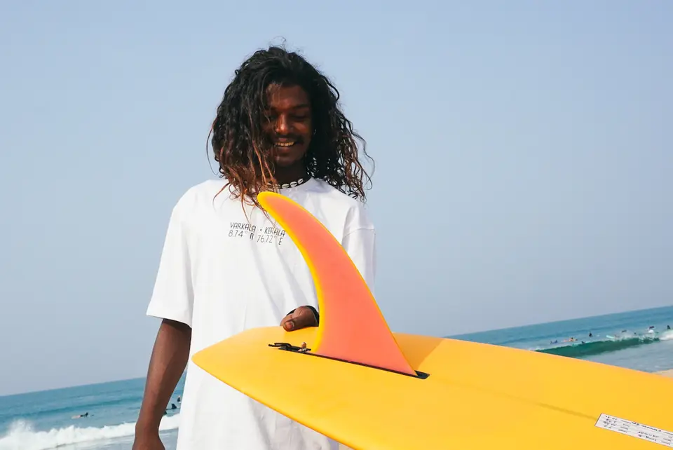 Kuma, smiling, holding a yellow surfboard with an orange fin on the beach