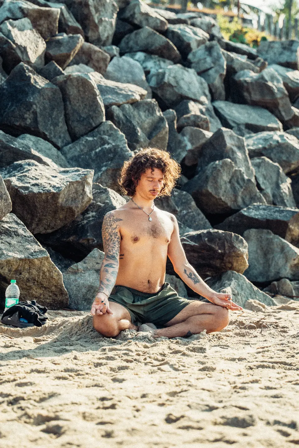 A man with curly hair sitting cross-legged in meditation on the sand, eyes closed, in front of dark boulders